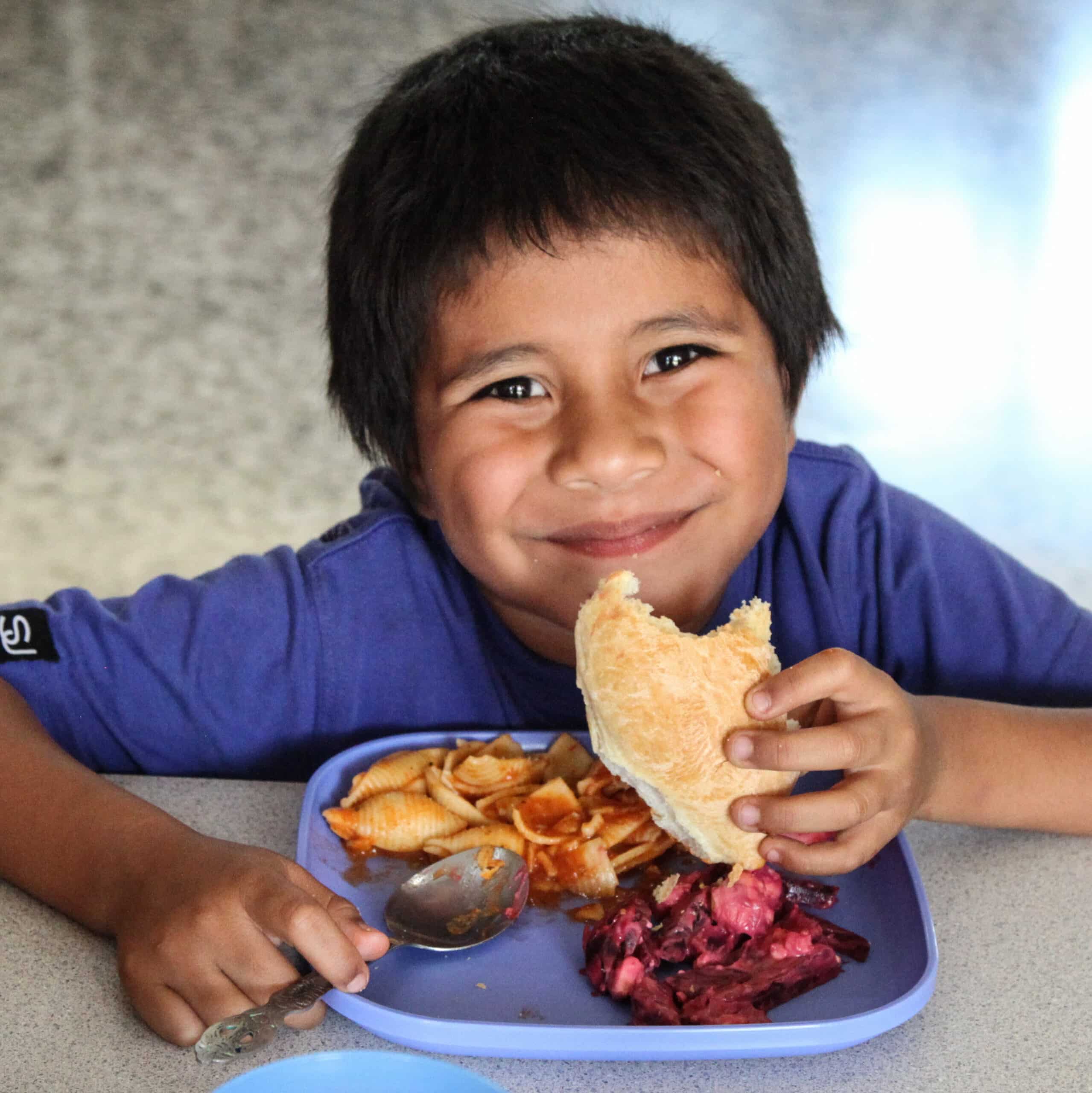 Der kleine Bub aus dem NPH Guatemala Projekt sitzt lächelnd vor einem vollen Teller mit Pasta und hält ein Brot in händen.